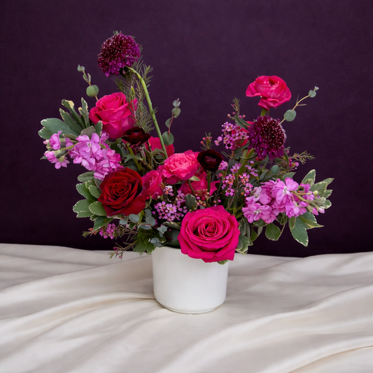 Bouquet of pink, red, and purple flowers in a white vase on a white surface with a dark purple background