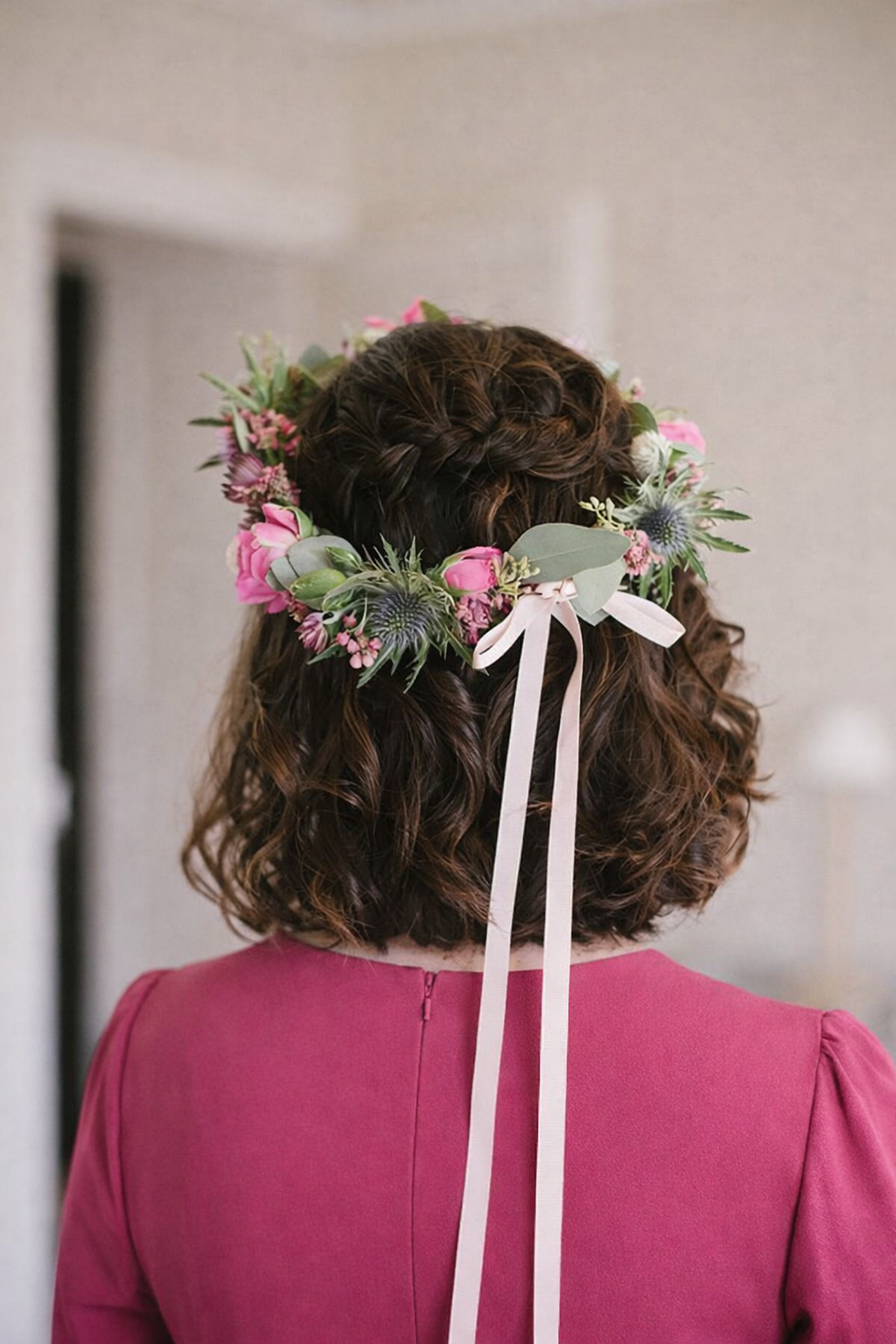 Person wearing a floral headpiece with pink flowers and green leaves, standing against a neutral background.
