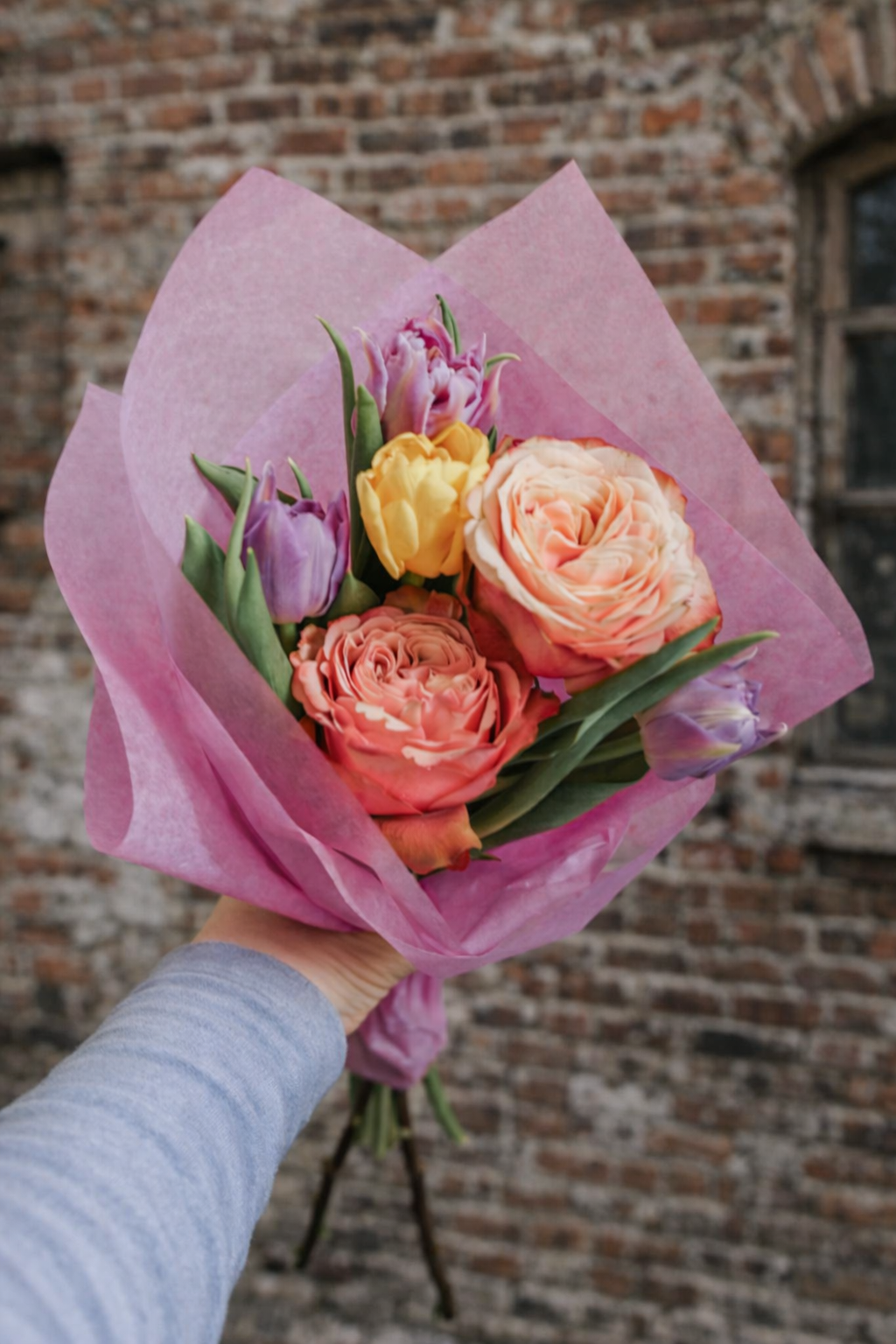 Bouquet of flowers wrapped in pink paper held against a brick wall.