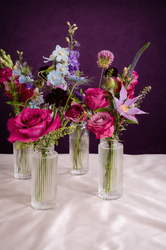 Four glass vases with colorful floral arrangements on a white surface with a purple background