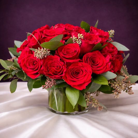 Bouquet of red roses in a clear vase on a white surface with a purple background