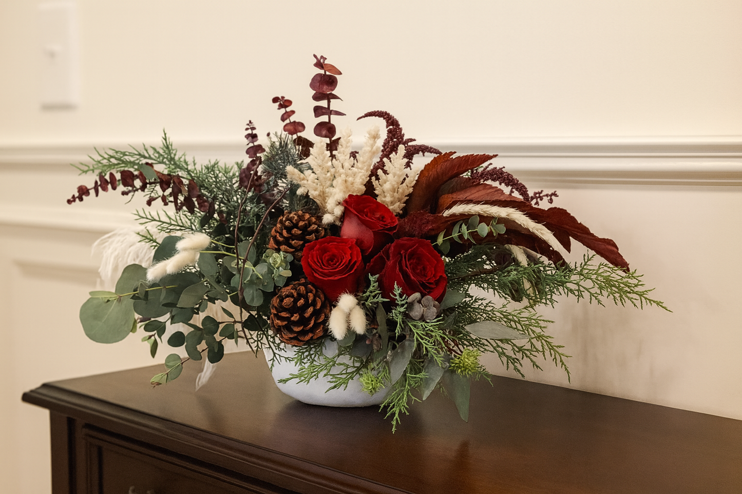 Floral arrangement with red roses, greenery, and pinecones in a white vase on a wooden surface.