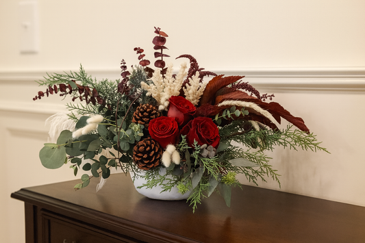 Floral arrangement with red roses, greenery, and pinecones in a white vase on a wooden surface.